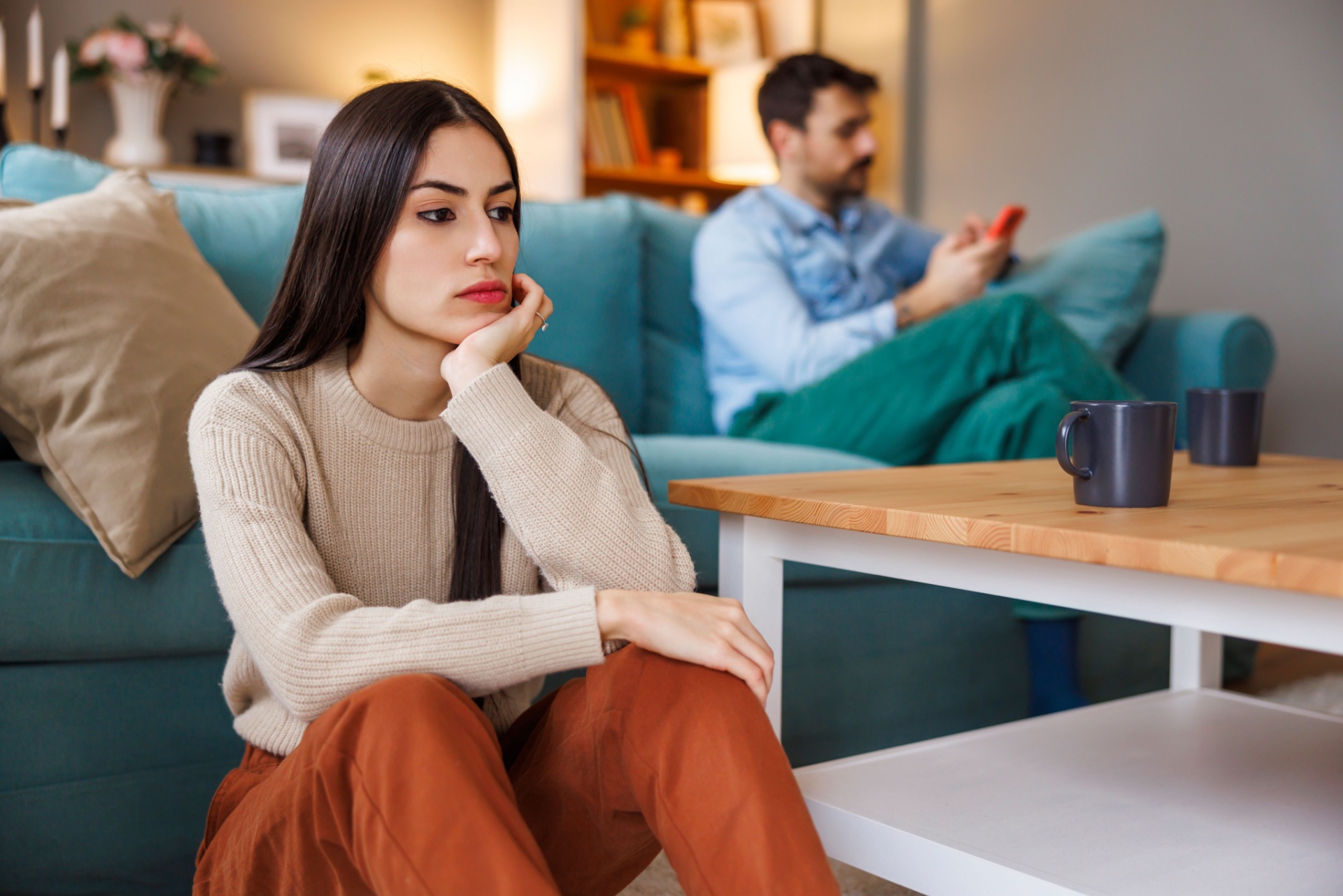 Kvinne som sitter på gulvet med ryggen mot sofaen hvor mannen hennes sitter og tekster på mobilen, ignorerer partneren sin. Partner er lei seg. Samlivsproblemer