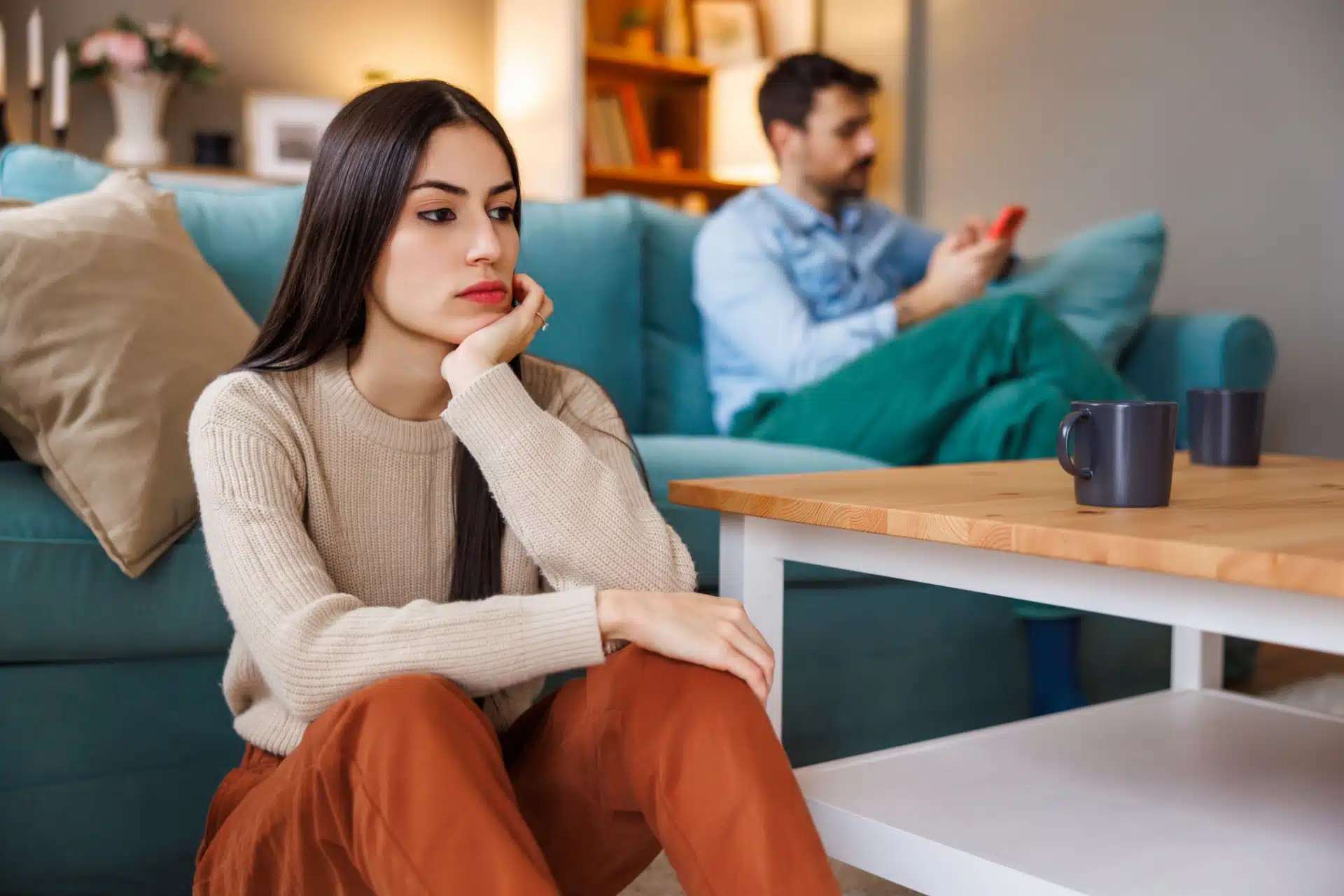 Kvinne som sitter på gulvet med ryggen mot sofaen hvor mannen hennes sitter og tekster på mobilen, ignorerer partneren sin. Partner er lei seg. Samlivsproblemer